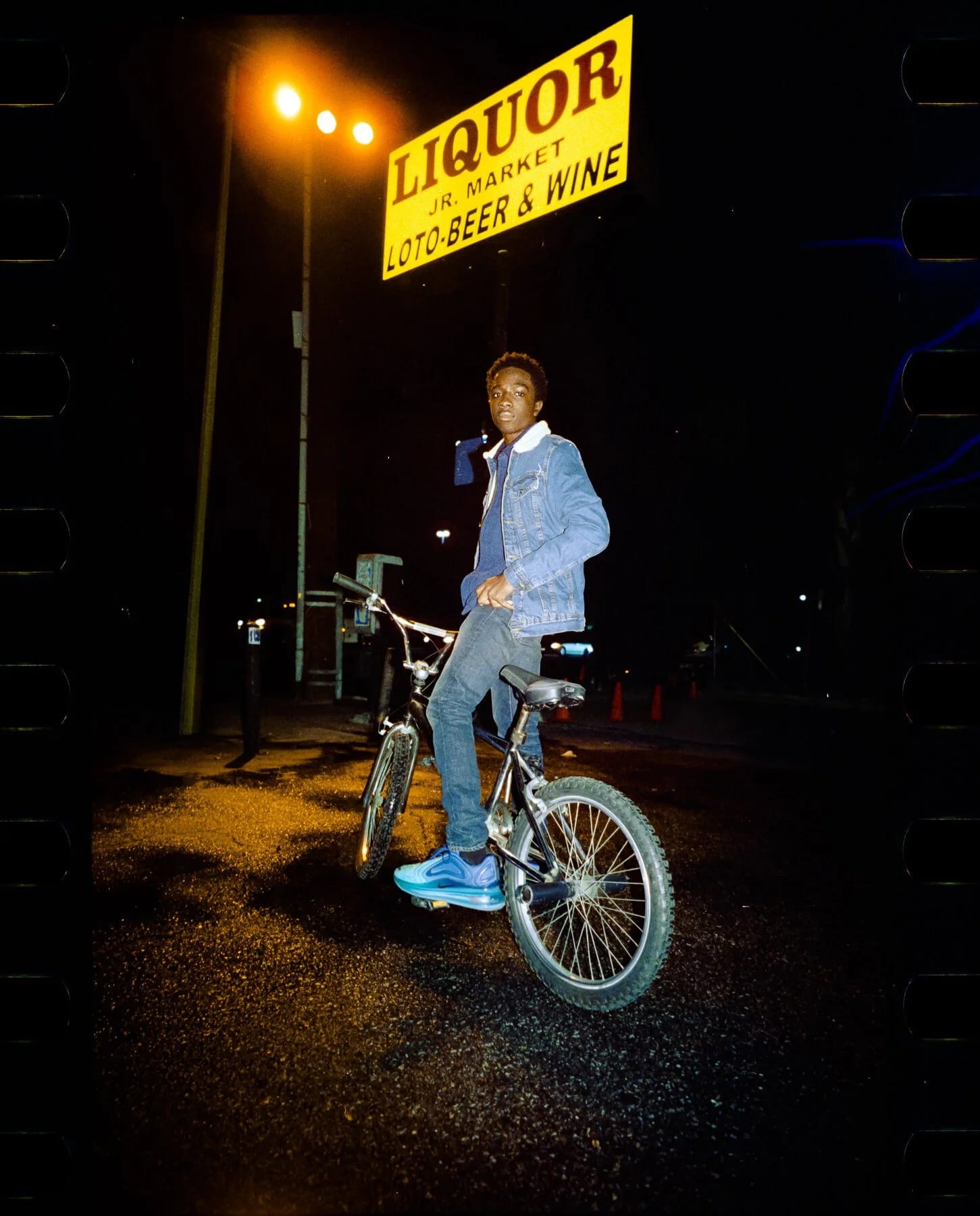 A young man in a denim jacket sitting on a bicycle under a yellow Liquor sign at night, lit by flash.