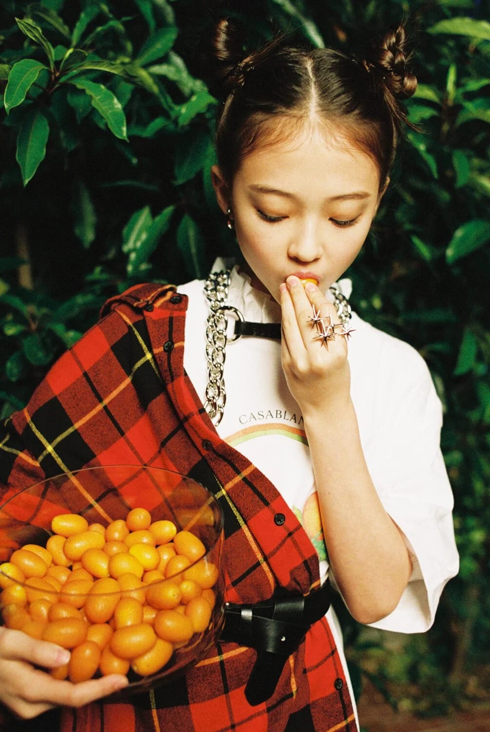 A young woman with hair buns in a red tartan shirt eating from a bowl of kumquats among green leaves.