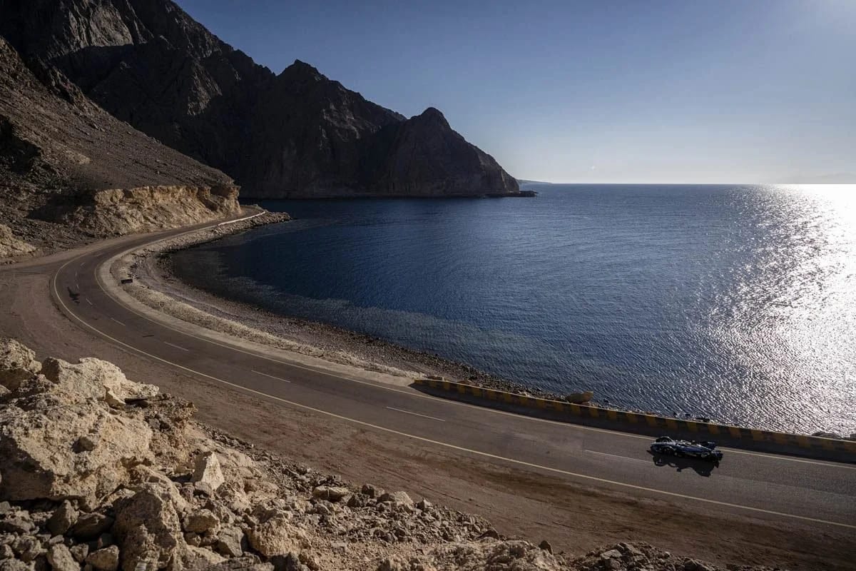 A coastal road curving around a bay below dark cliffs, a car driving along it in the sun.