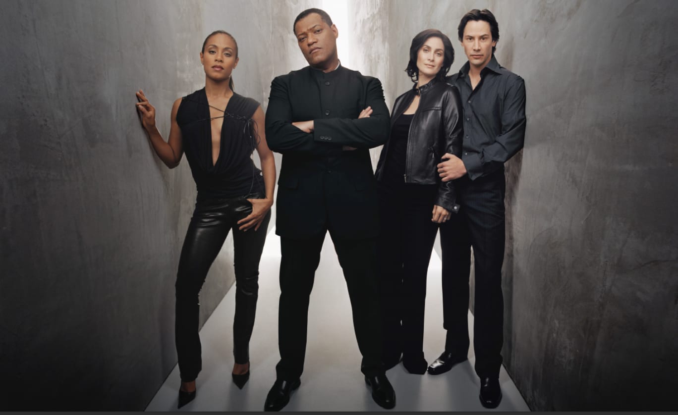 Four actors in black leather standing side by side against a grey wall, one woman with her arm raised.