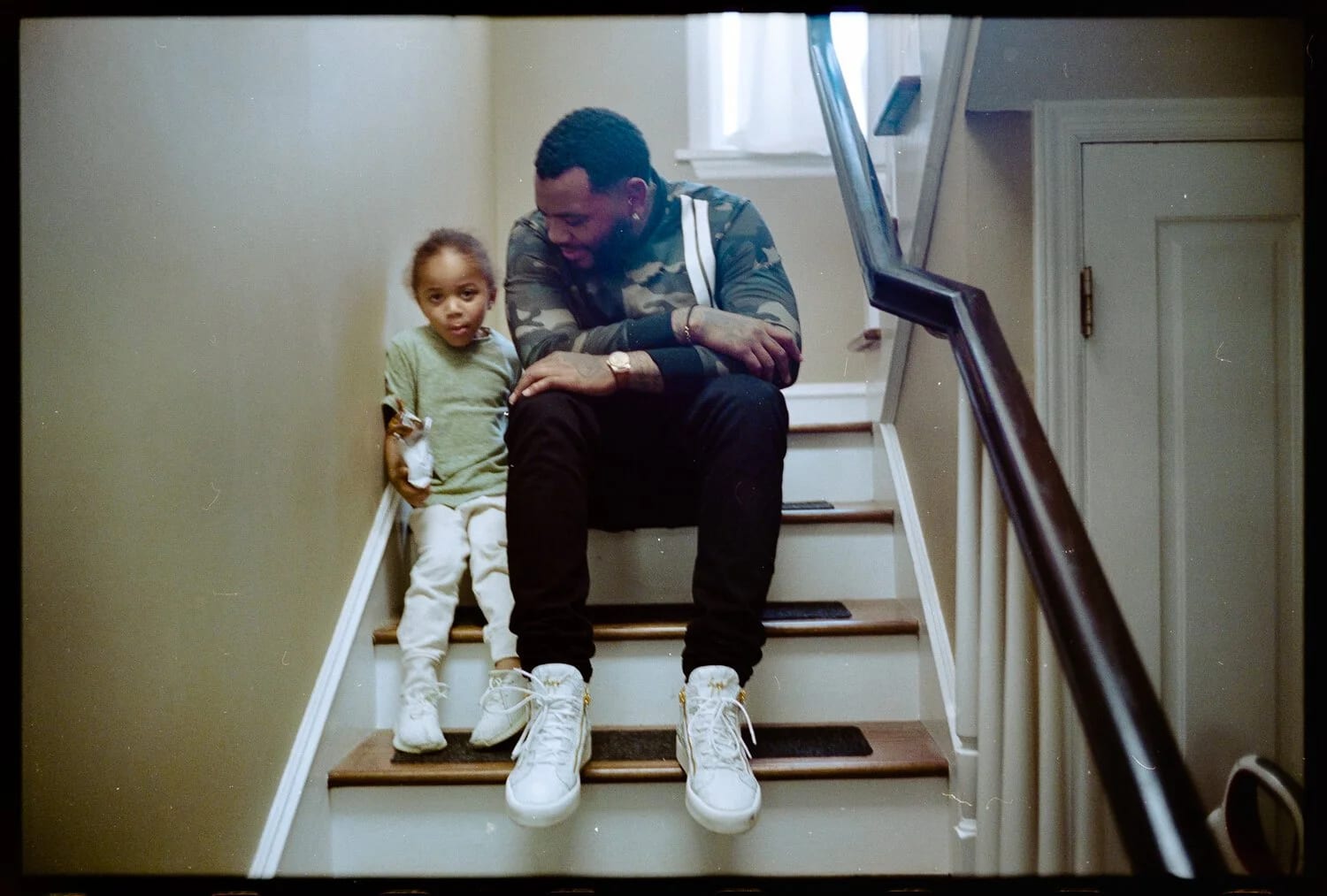 A man in a camouflage jacket sitting on a staircase with a small child leaning against him, both in white trainers.