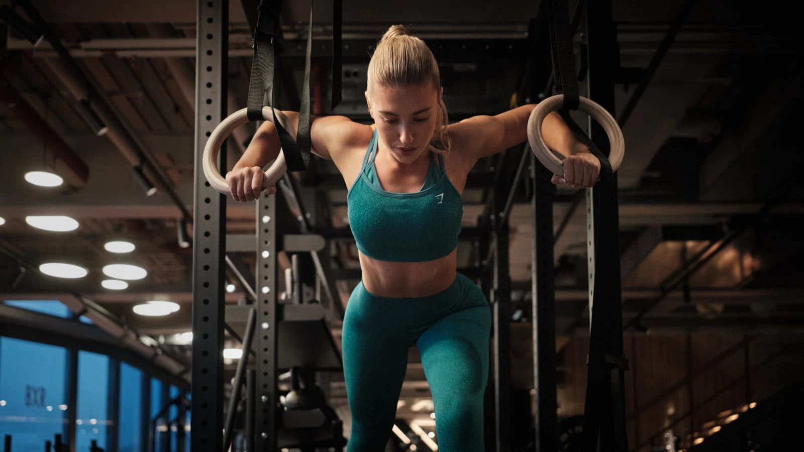 A woman in teal sportswear hanging from gymnastic rings in a dark gym.