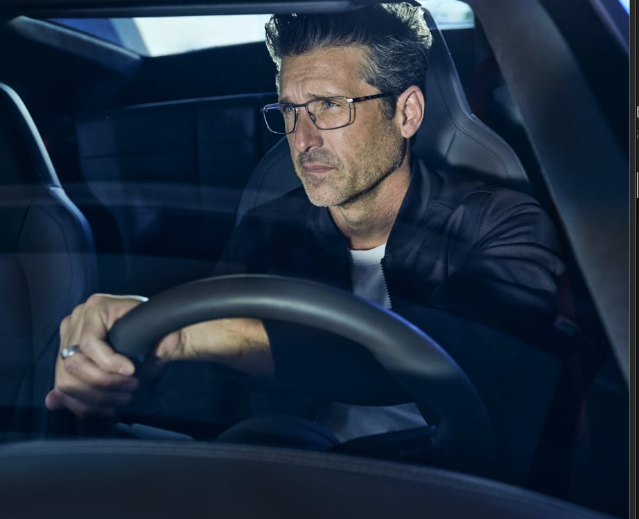 A man in glasses at the wheel of a car, seen through the windscreen.