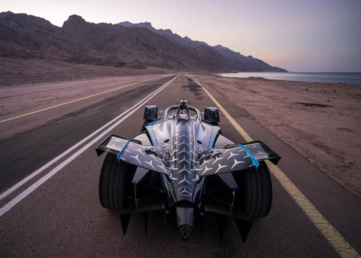 A black and silver open-wheel racing car parked on an empty coastal road, mountains and sea beyond.
