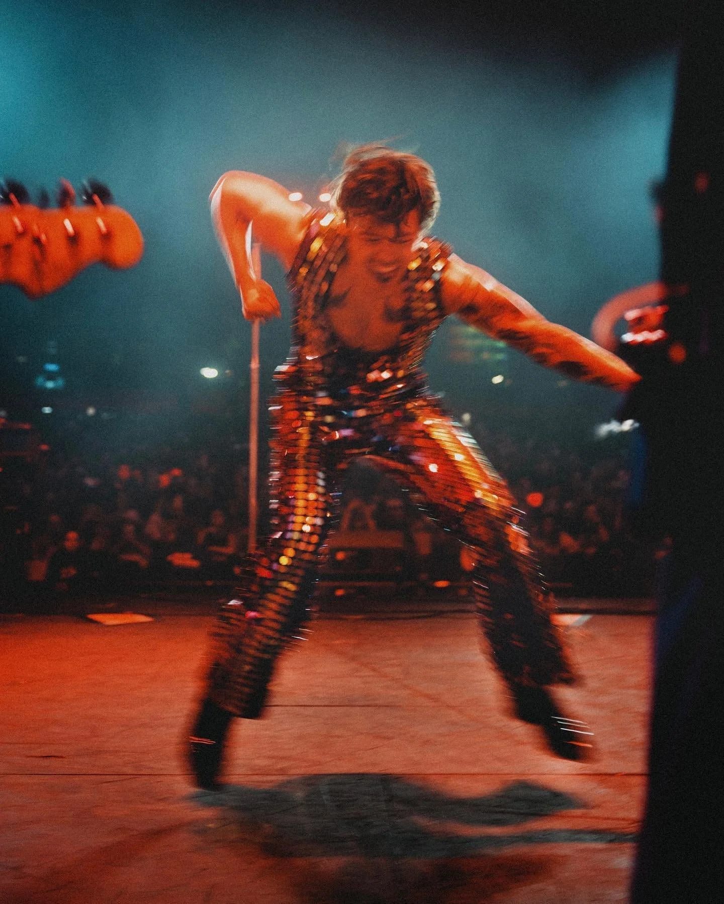 A performer in a mirrored sequinned suit leaning into a microphone stand on stage, a crowd of lights beyond.