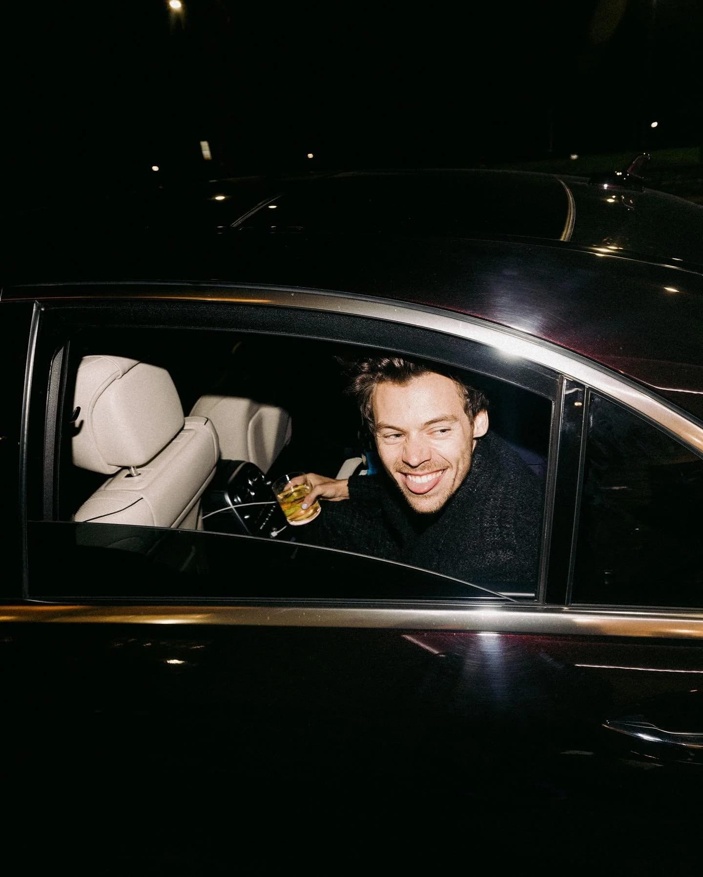 Harry Styles grinning out of the rear window of a dark car at night, a drink in his hand.