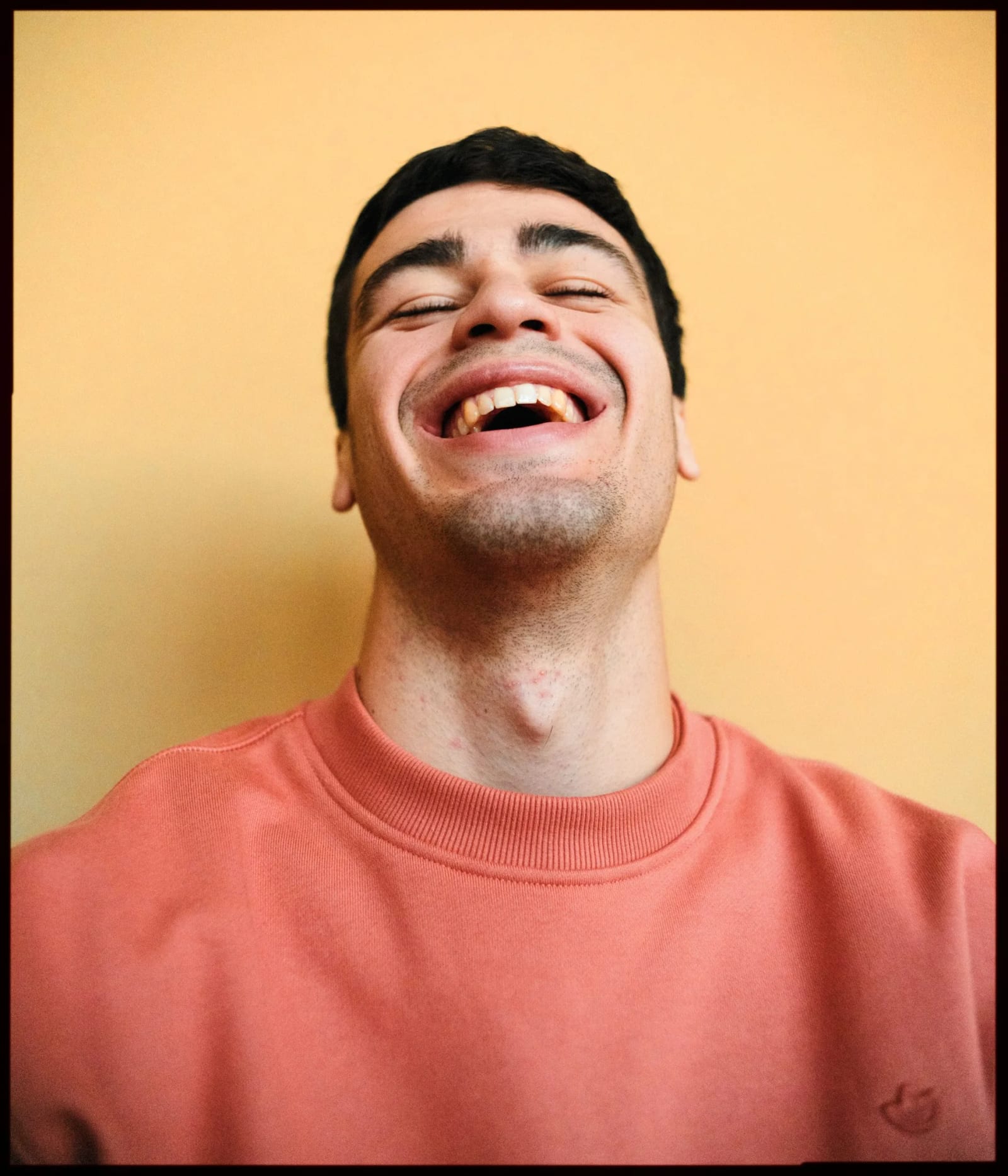 A young man in a coral sweatshirt laughing with his head tipped back against an orange-yellow wall.