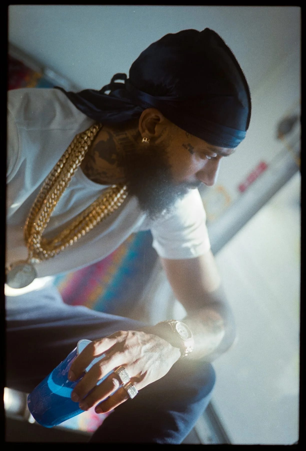 A man in a black durag and gold chains looking down, a bottle of blue drink in his hand, sunlight behind him.