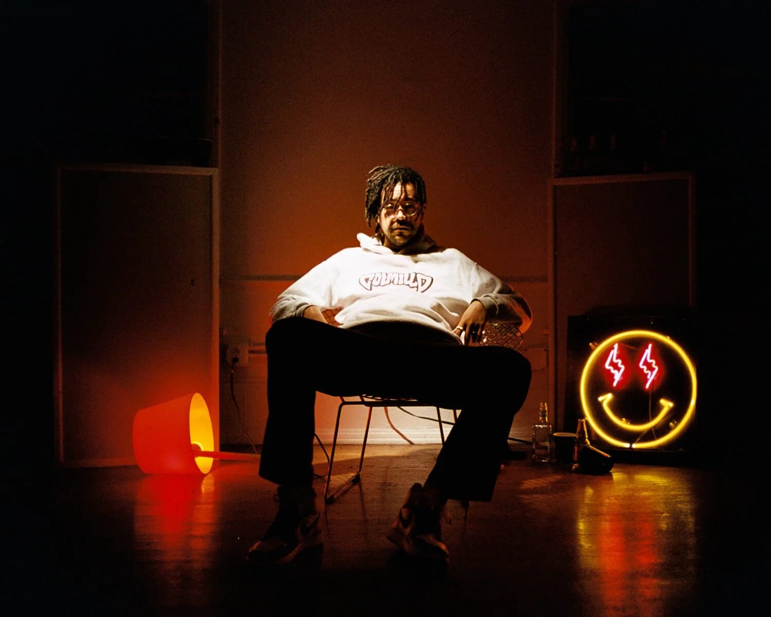 A man in a white sweatshirt sitting on a chair in a dark room with an orange lamp and a neon smiley face with lightning-bolt eyes.