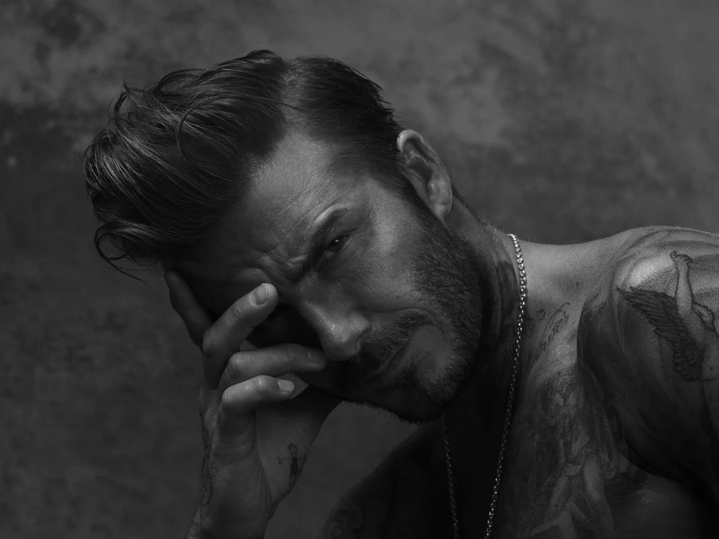 Black and white portrait of a tattooed man with slicked-back hair resting his head against his hand.