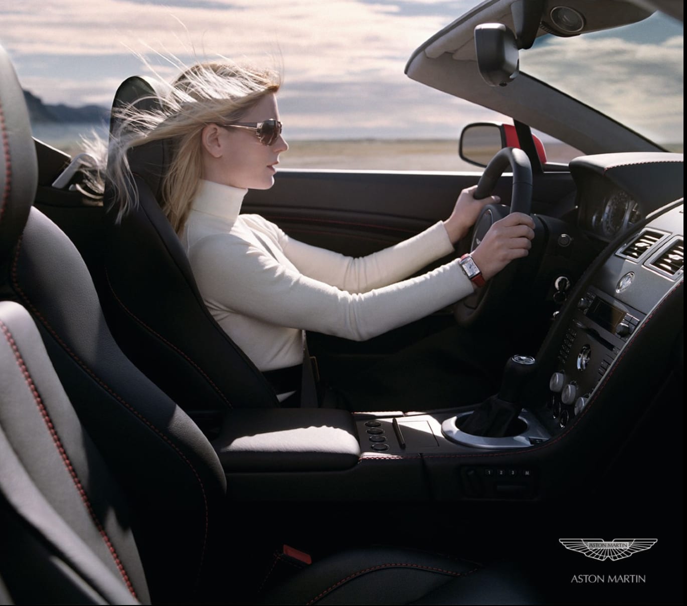 Aston Martin advertisement: a woman in sunglasses and a white turtleneck driving a convertible, her hair blown back.