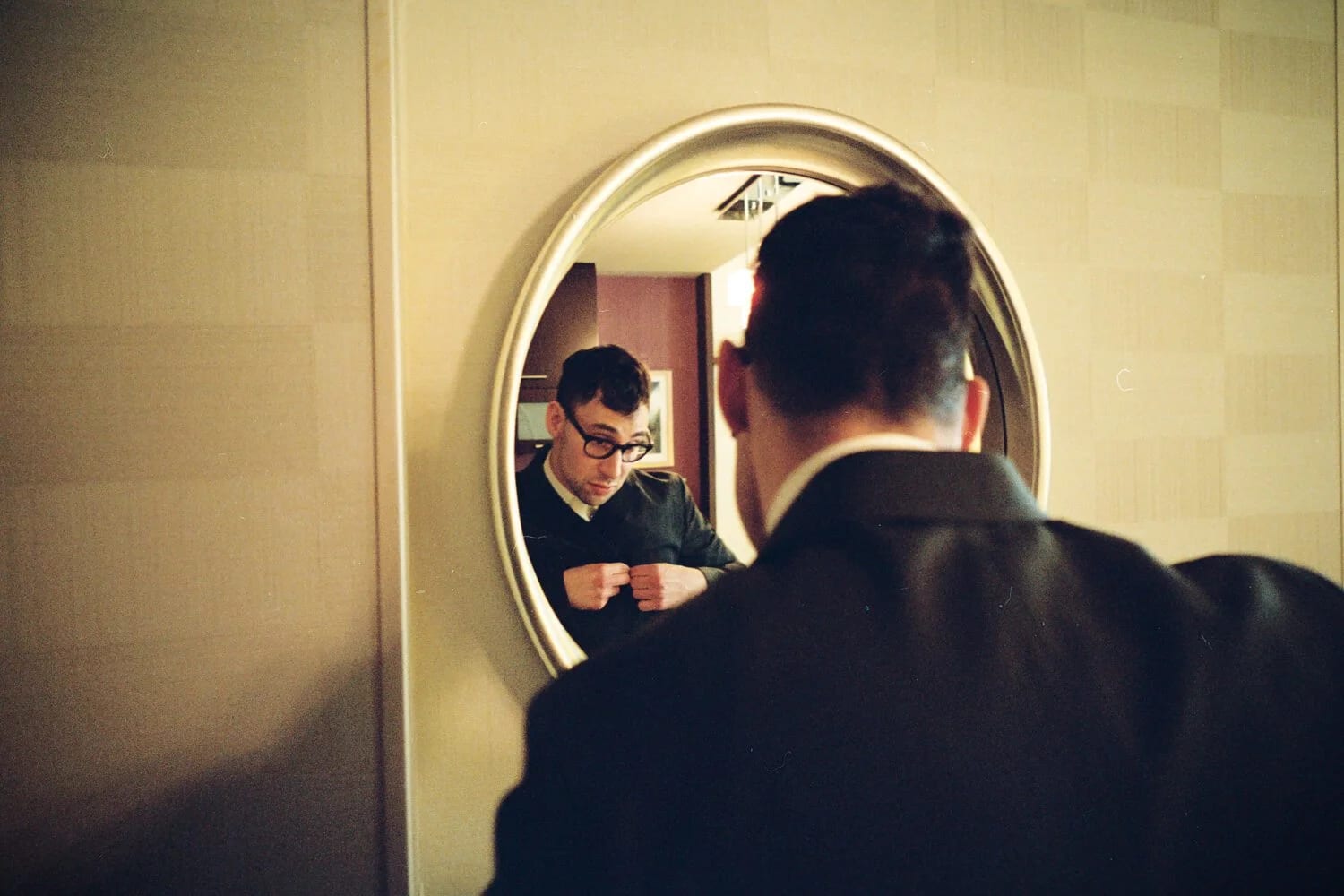 A man in a dark jacket seen from behind, his reflection in a round mirror showing glasses and a raised fist.