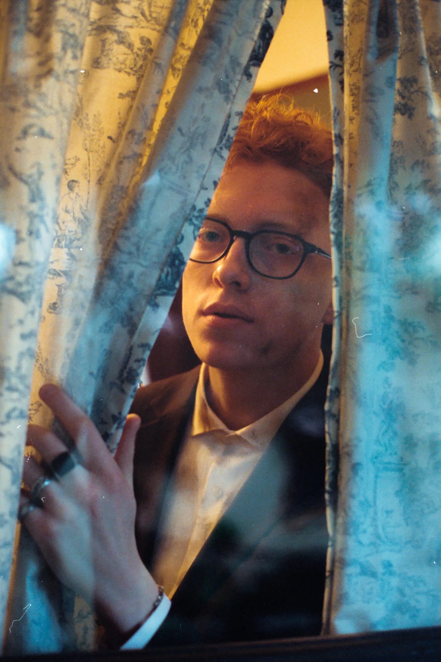A red-haired man in glasses and a suit peering through a gap in patterned curtains.