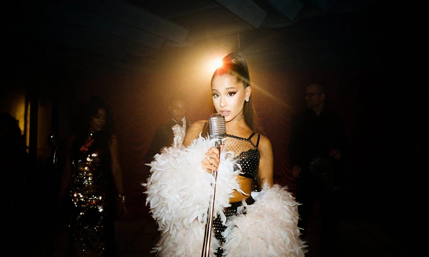 A woman in a white feathered coat and a high ponytail at a vintage microphone under a single spotlight.
