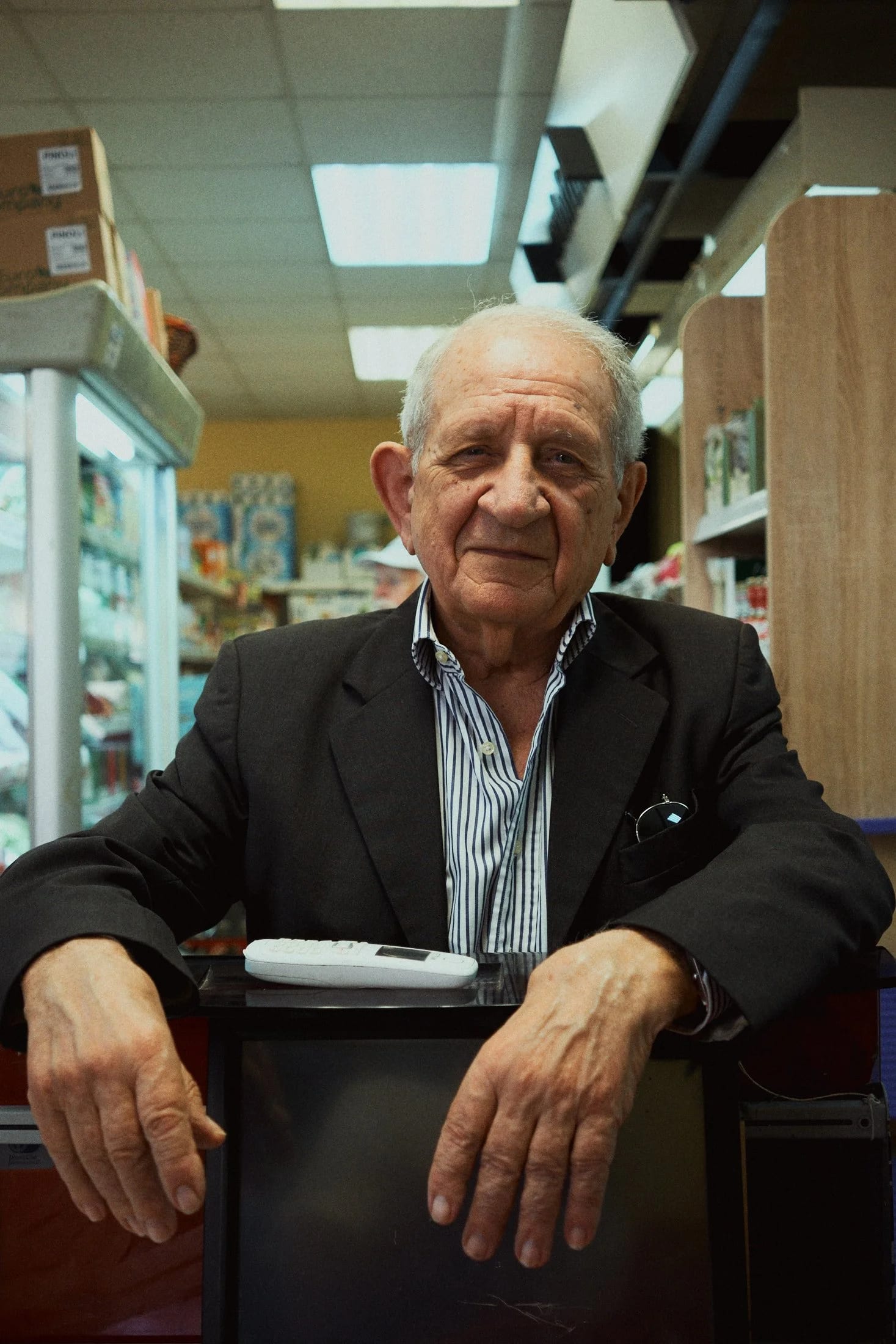 An older man in a striped shirt and dark jacket resting his hands on a shop counter beneath fluorescent lights.