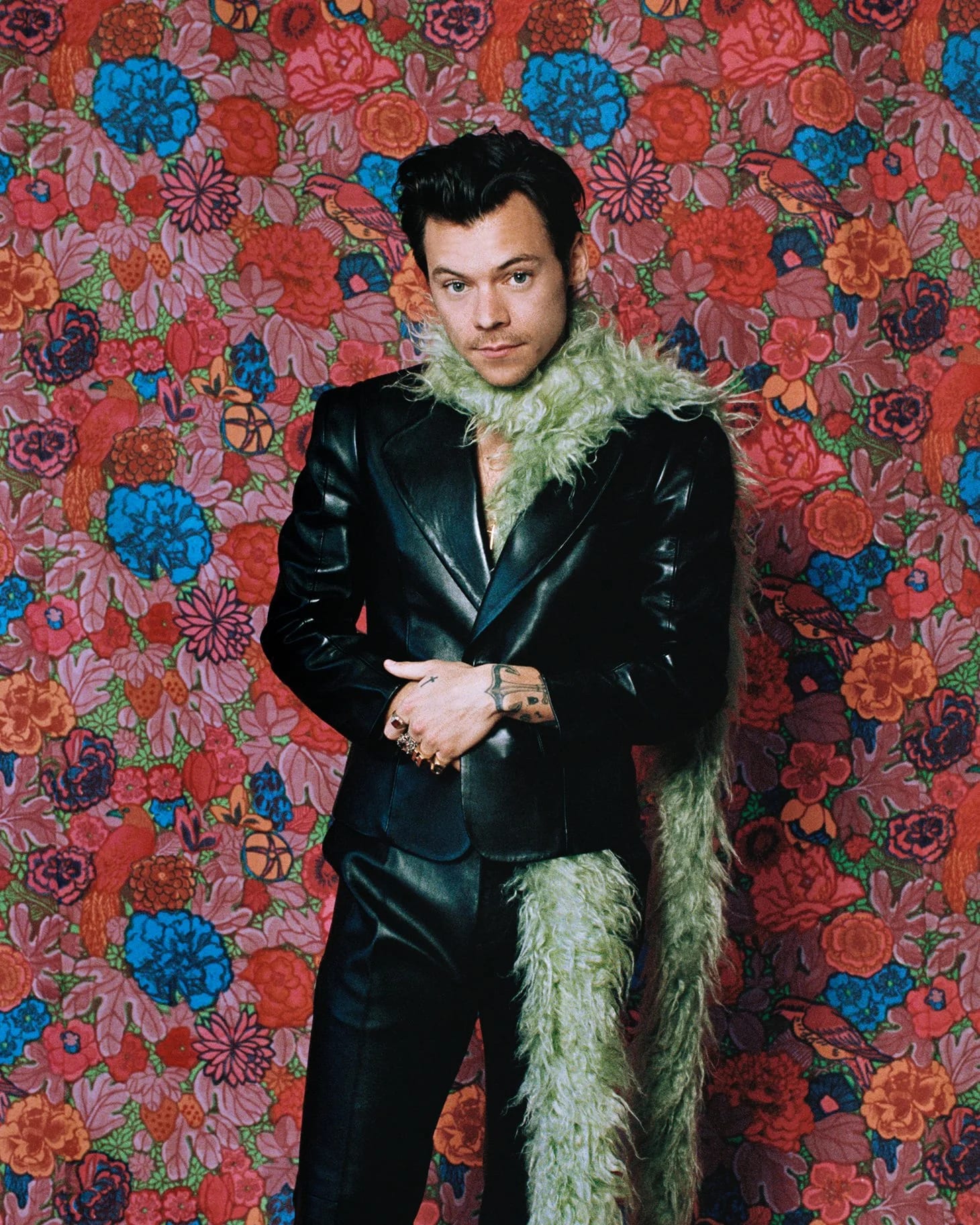 Harry Styles in a black leather suit and green feather boa against a red floral wallpaper.