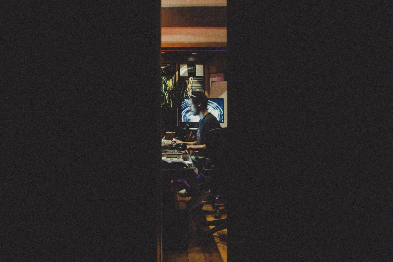 A man at a studio desk seen through a narrow doorway in the dark.