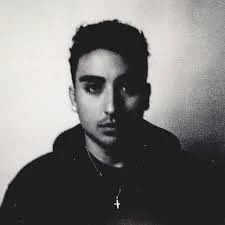 Grainy black and white portrait of Chris Villa in a dark hoodie with a cross pendant, looking into the camera.