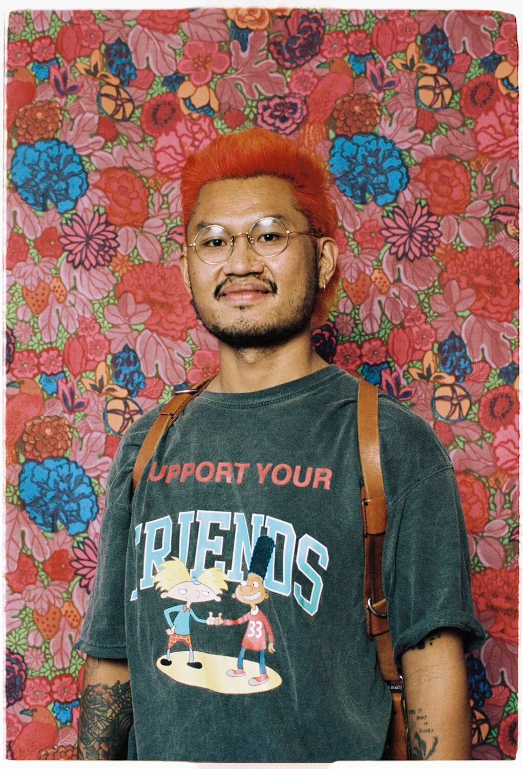 Anthony Pham with dyed orange hair and glasses in a Hey Arnold T-shirt reading Support your friends, standing against a floral wallpaper.