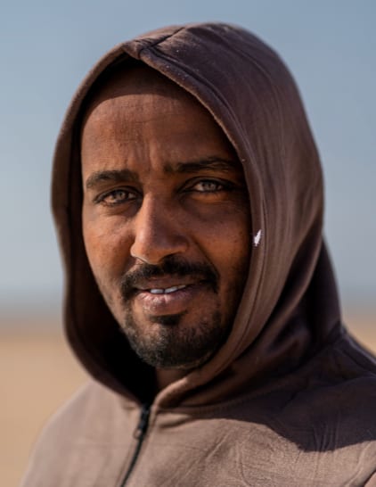 Close-up portrait of a smiling man in a brown hooded top, desert and sky behind him.