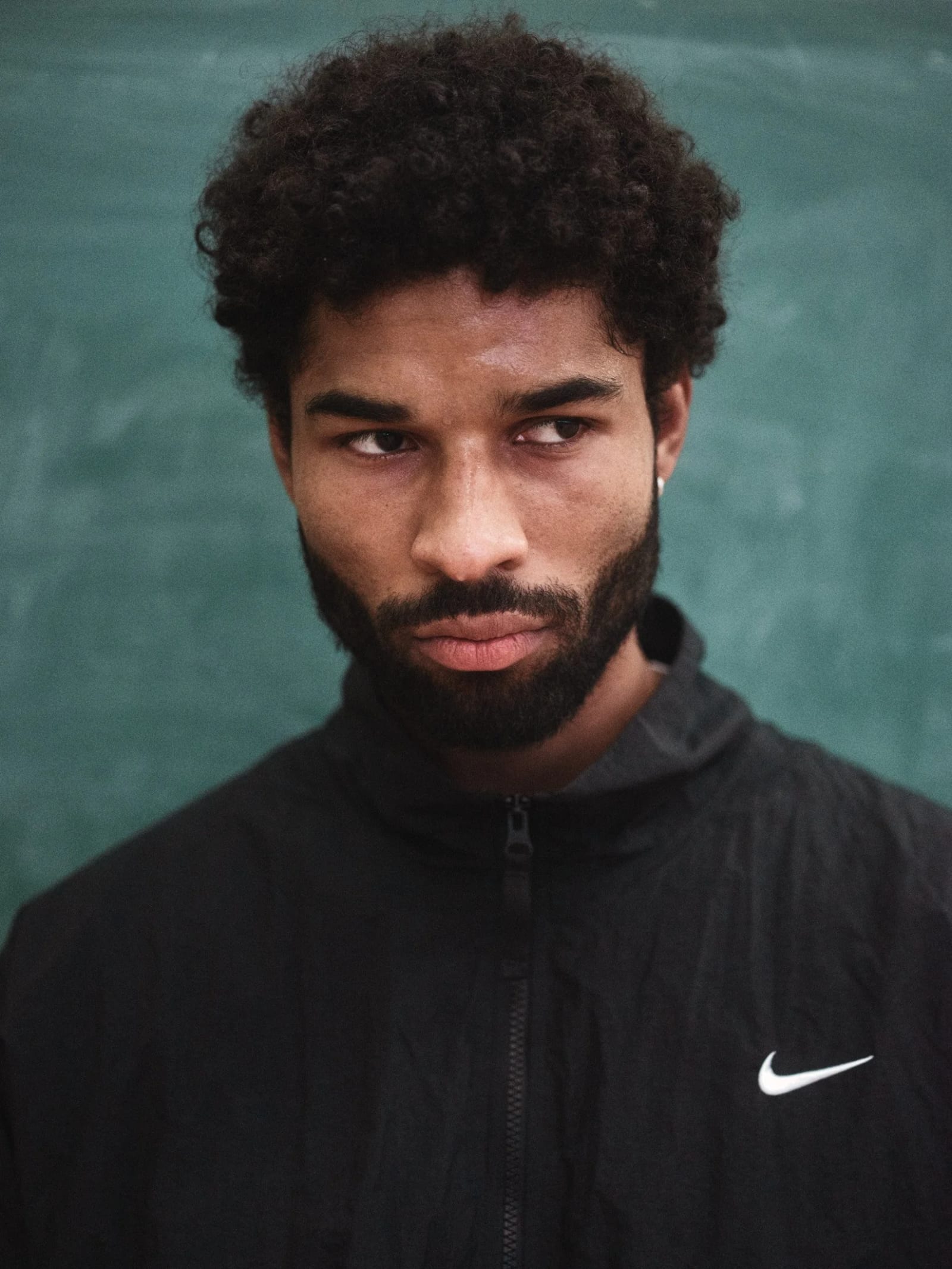 Portrait of a bearded man with curly hair in a black Nike jacket glancing sideways against a green wall.
