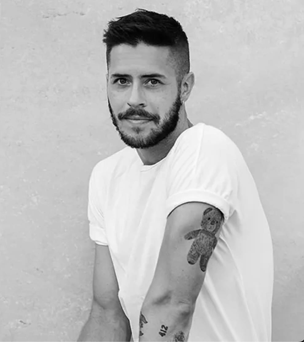 Black and white portrait of Jake Wilson in a white T-shirt with a teddy-bear tattoo on his arm, against a plain wall.