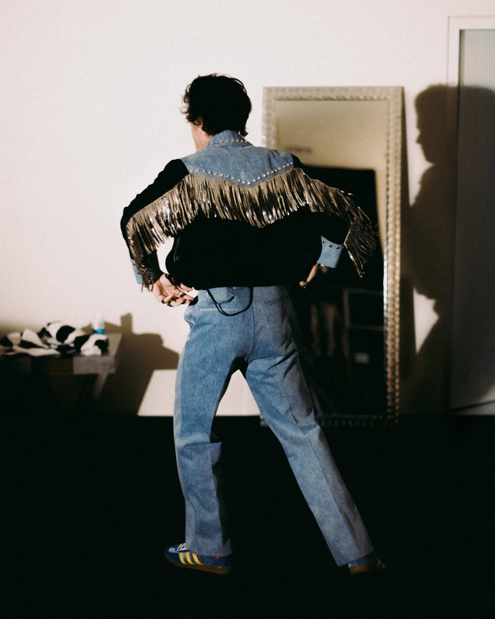 A man seen from behind in a fringed denim jacket and jeans walking toward a mirror in a bare room.