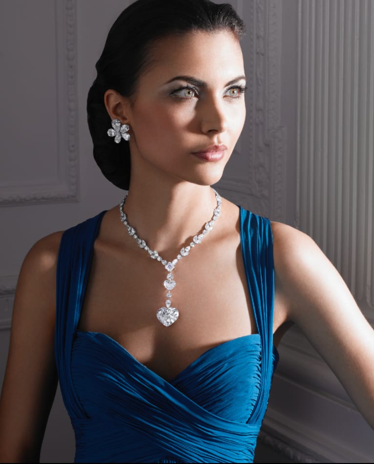 A dark-haired woman in a blue draped gown wearing a diamond necklace and earrings, looking off to the side.