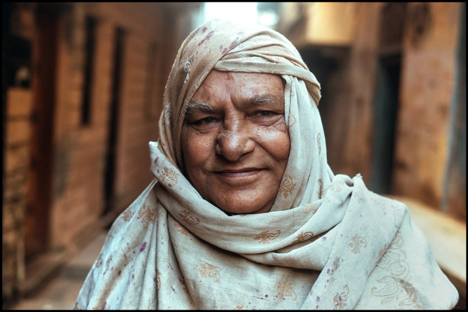 An older woman in a pale patterned headscarf smiling in a narrow sunlit alley.