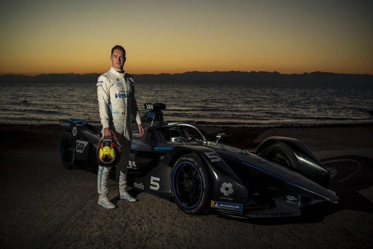 A racing driver in a white suit holding his helmet beside a black racing car on a beach at sunset.