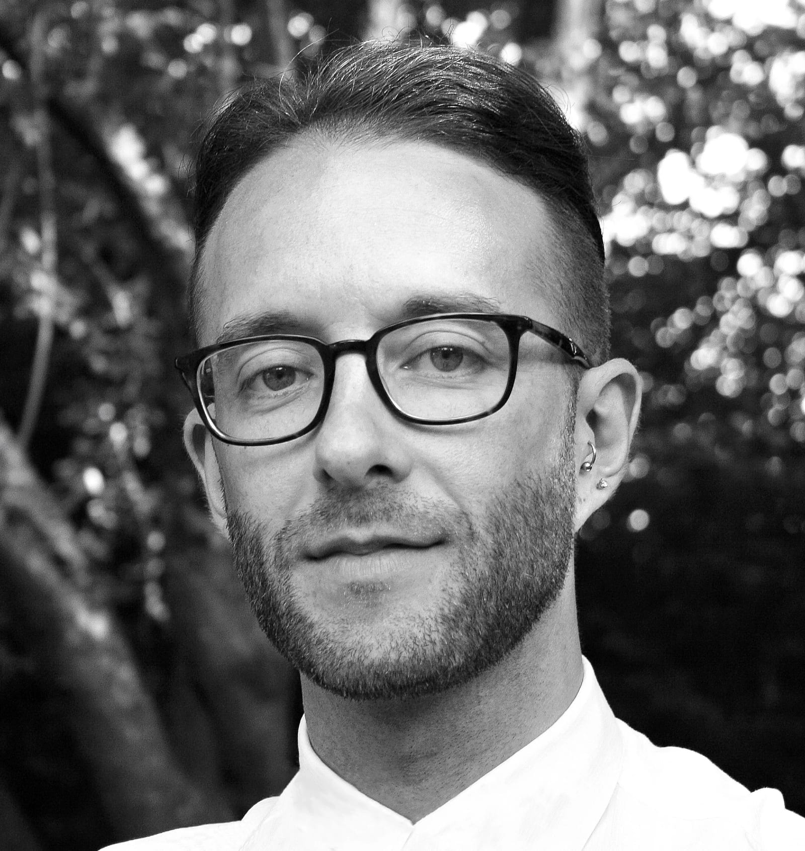 Black and white portrait of Florent Igla in glasses and a white shirt in front of sunlit leaves.