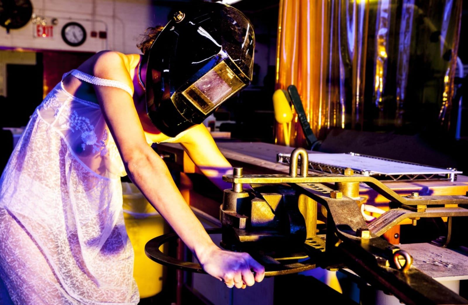 A woman in a sheer lilac dress and a welding mask leaning over a workbench in a purple-lit workshop.
