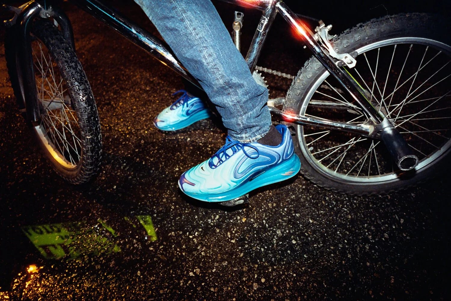 Close-up of blue and silver trainers and jeans standing astride a bicycle on wet asphalt at night.