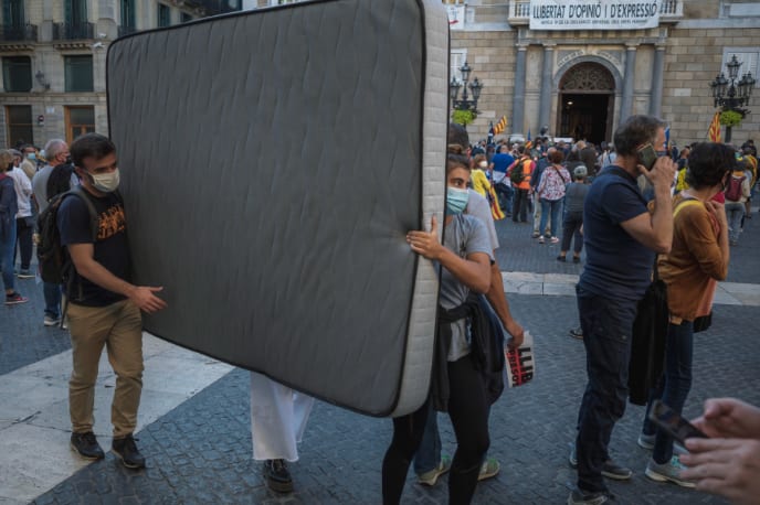 Two people in face masks carrying a mattress through a crowded city square.