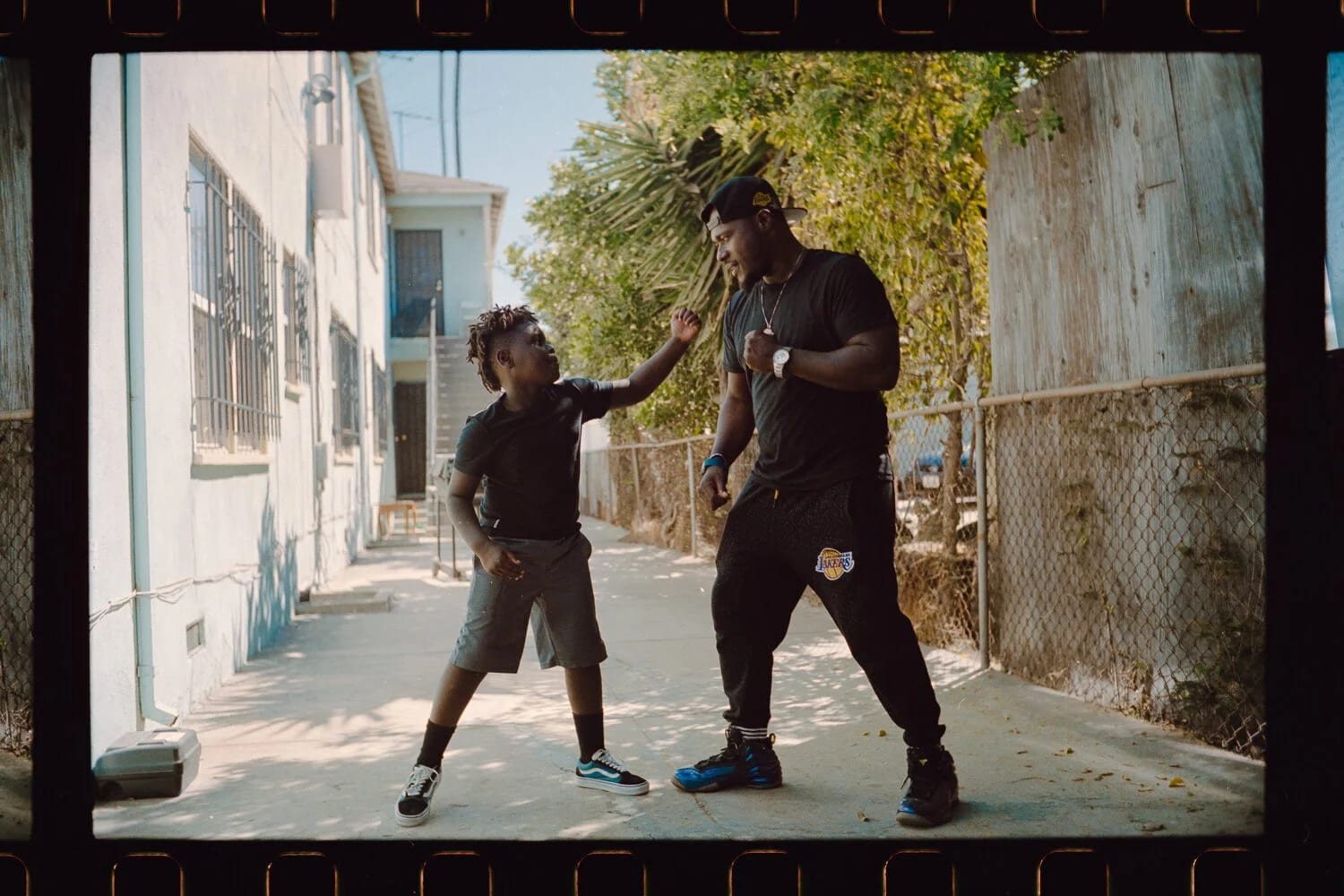 A film-strip frame of a young boy in black play-fighting with a man on a sunny street beside a chain-link fence.