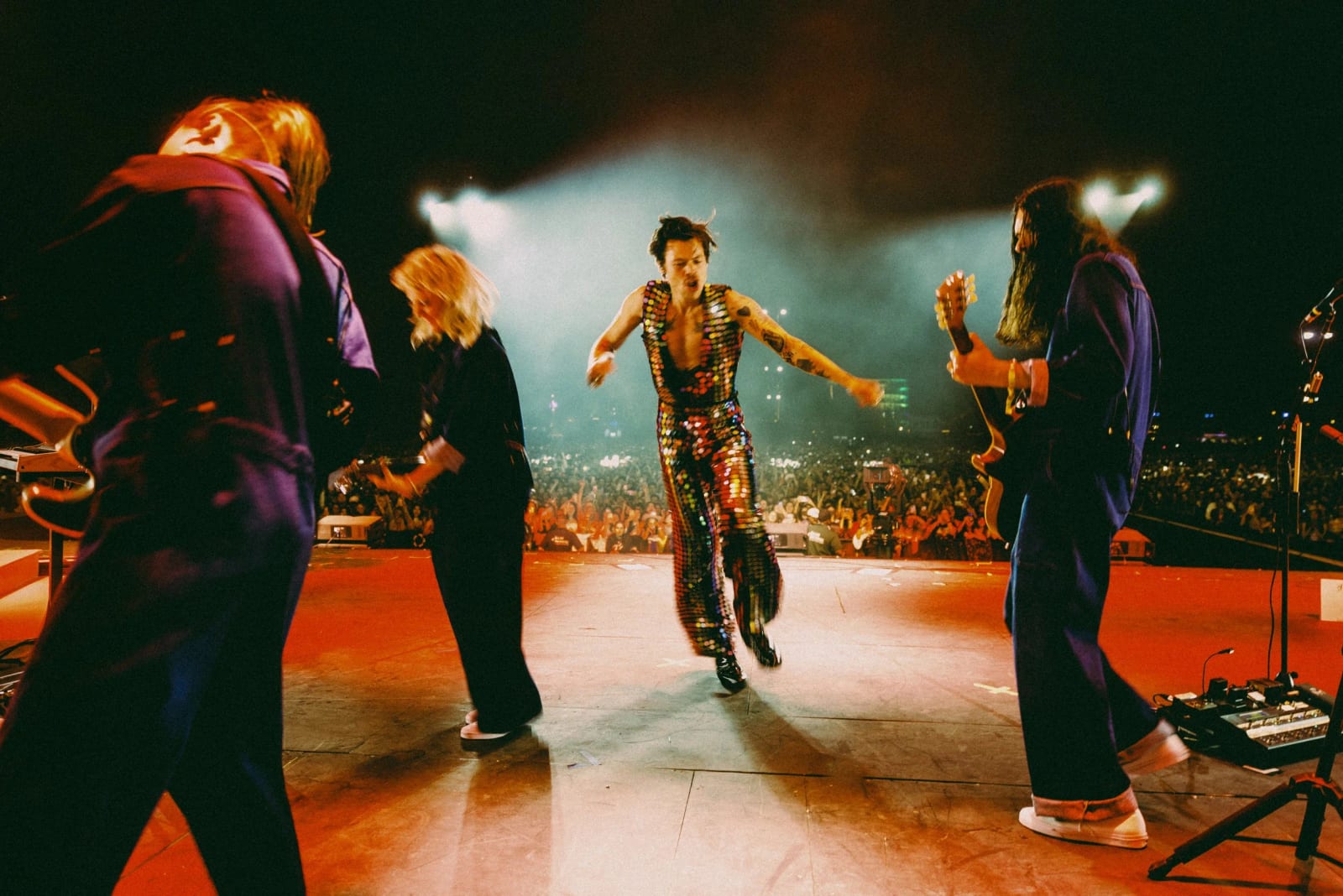 A singer in a sequinned suit running across a stage between musicians, a lit crowd stretching into the distance.
