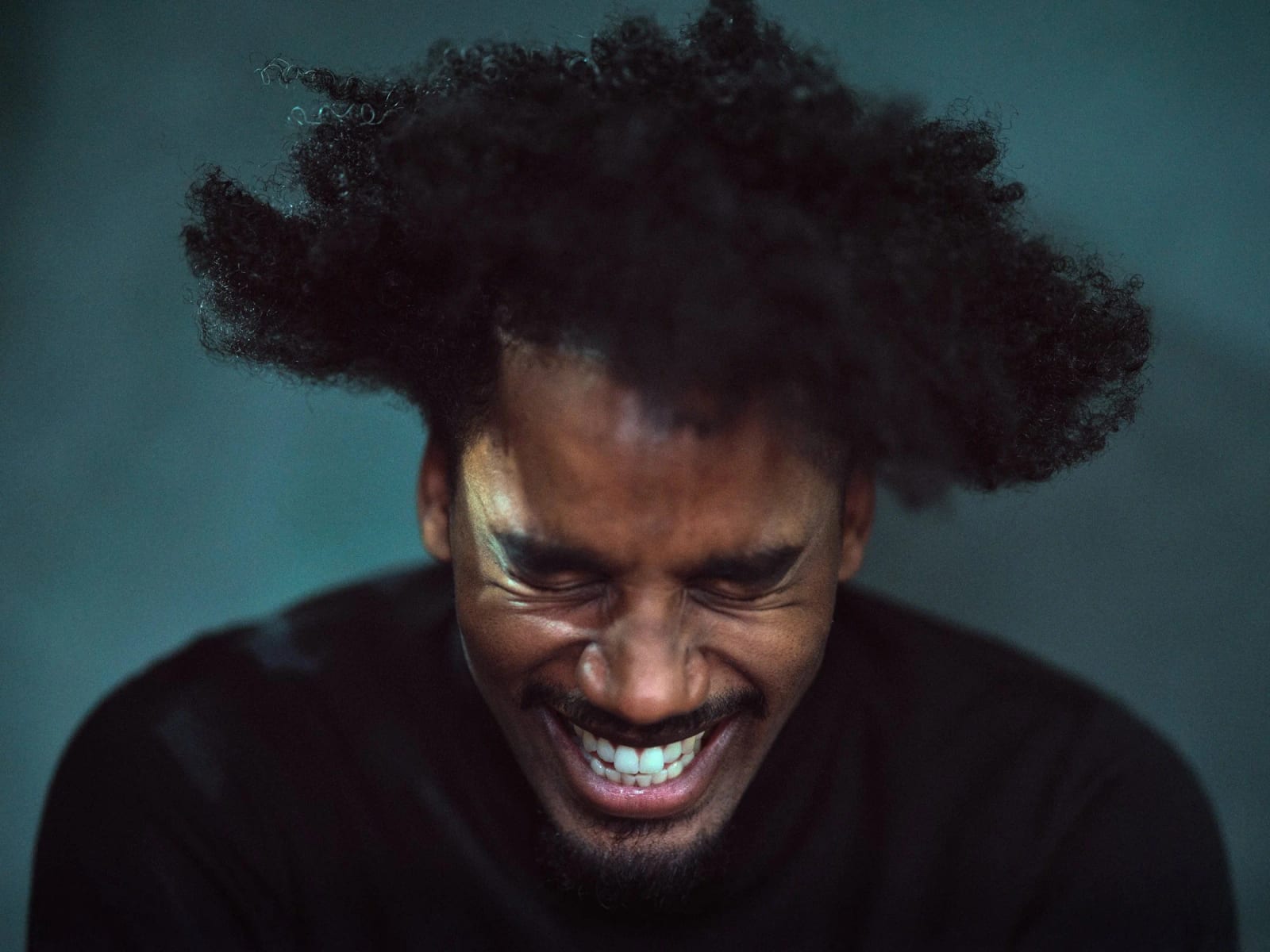 A man laughing with his eyes squeezed shut, his hair flung upward, lit in teal light against a dark background.