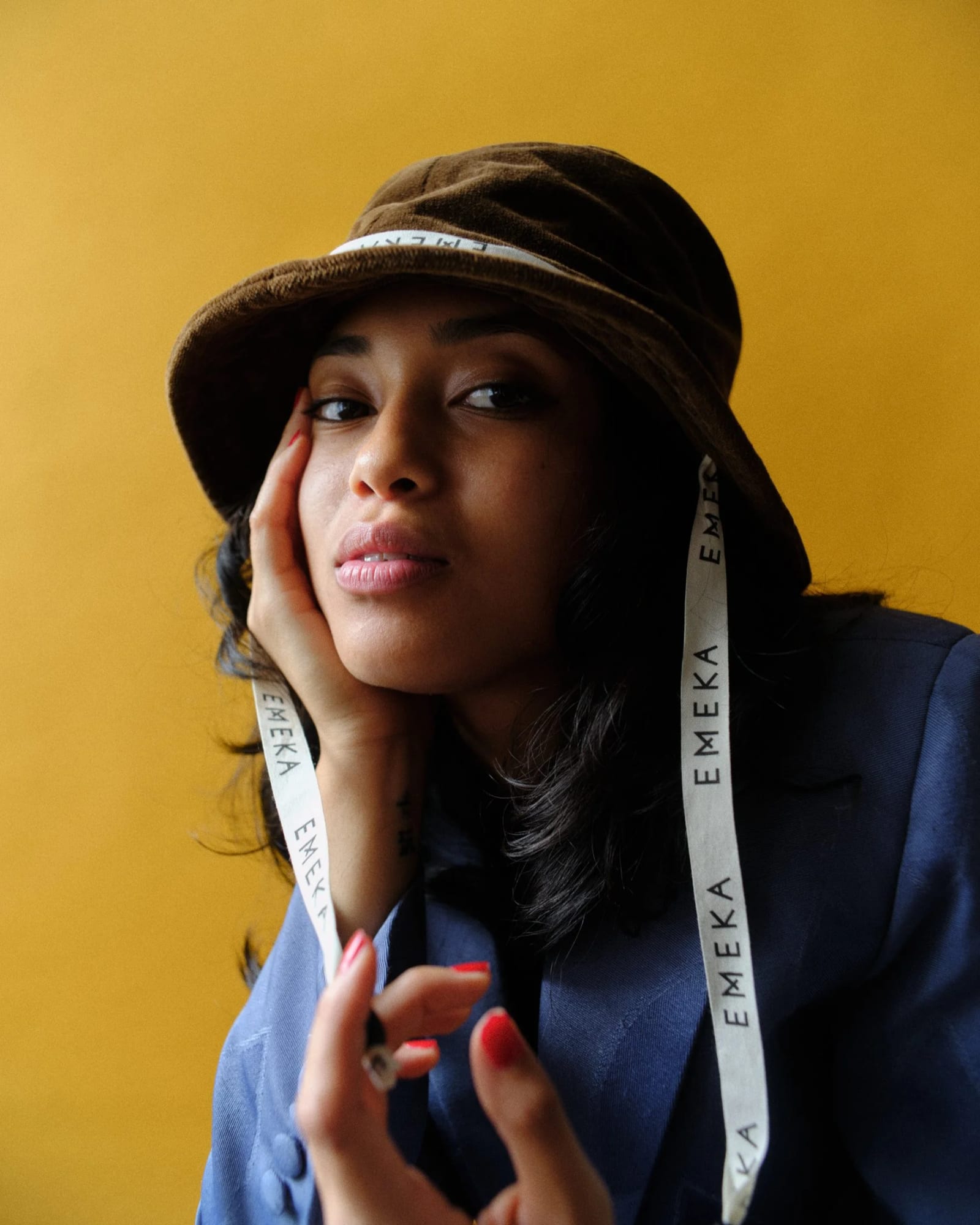A woman in a brown bucket hat with EMEKA straps and a blue jacket, her hand against her cheek, against a yellow wall.