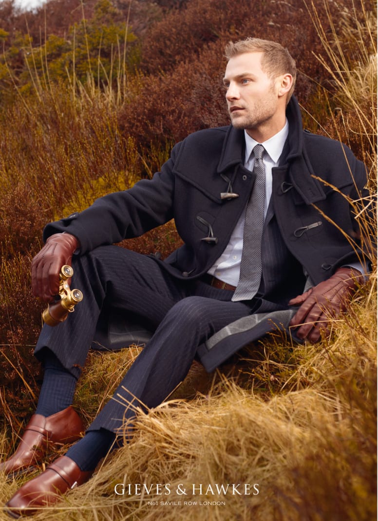 Gieves and Hawkes advertisement: a man in a navy coat and pinstripe suit sitting in tall autumn grass with a hip flask.