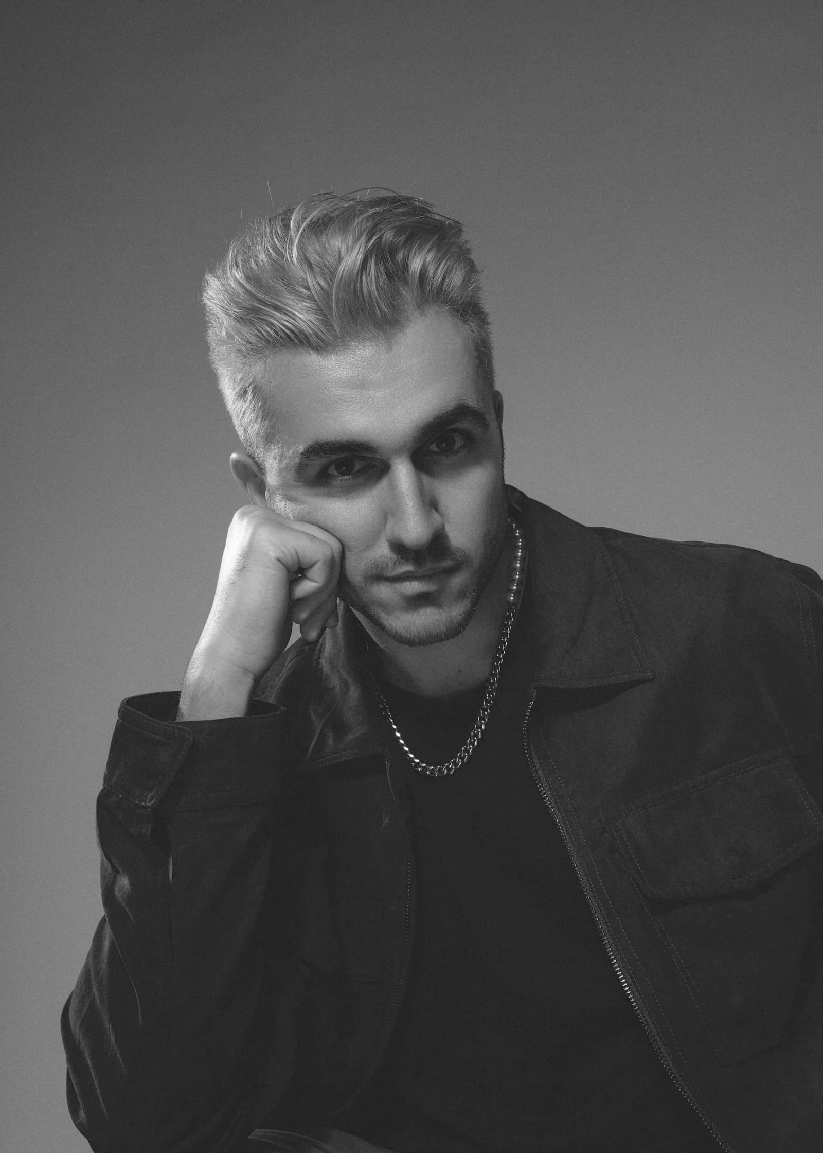 Black and white portrait of Arrad with swept-back hair and a chain, resting his cheek on his fist.