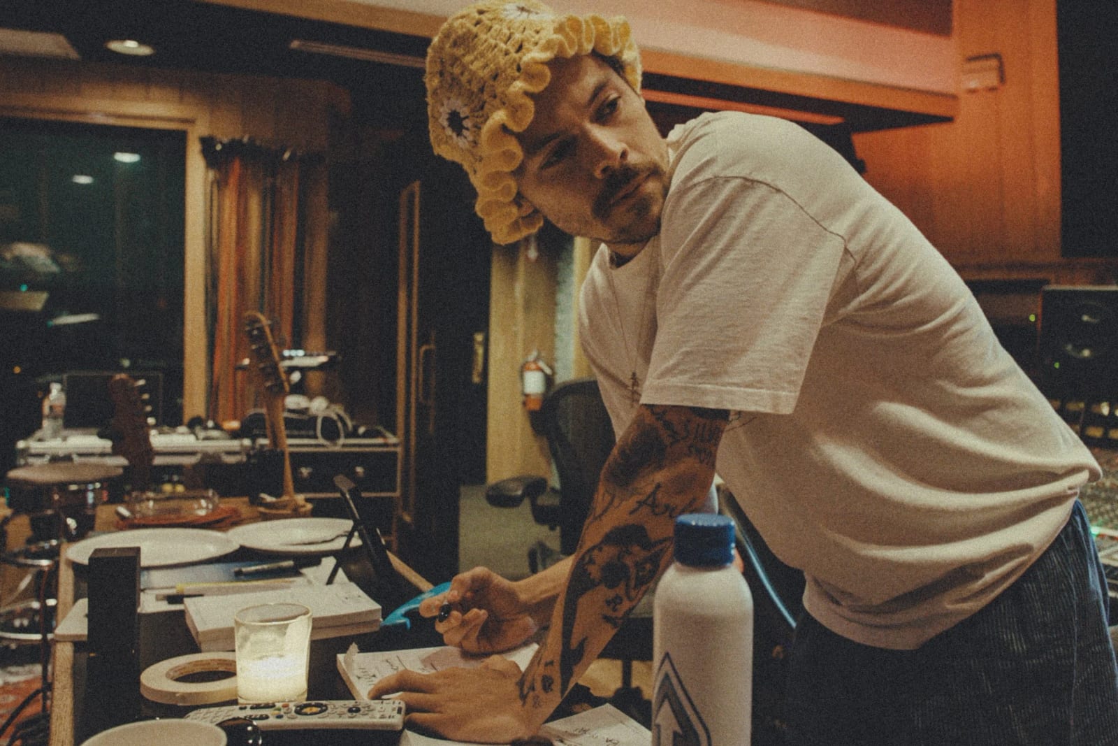 Harry Styles in a curly blonde wig and white T-shirt leaning over a studio table to write a note.