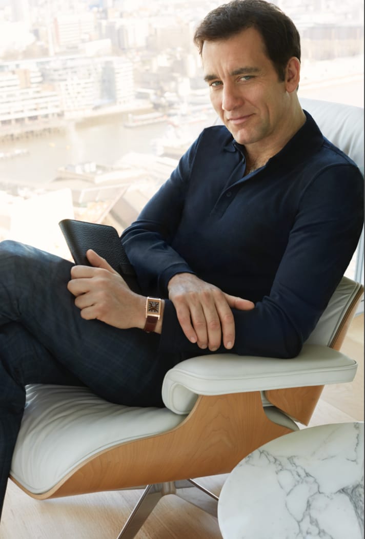 A man in a navy top sitting in a leather lounge chair with a tablet, a city skyline through the window.