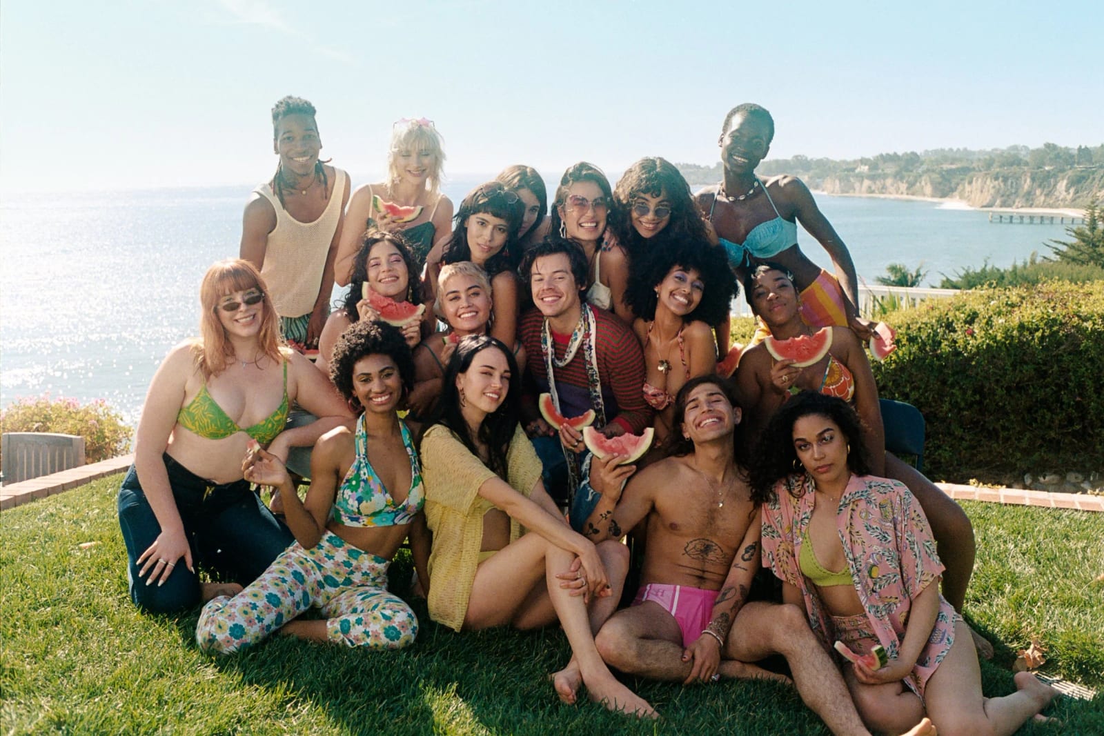 A group of about twenty people in swimwear and summer clothes posing on a lawn above the sea, several holding slices of watermelon.