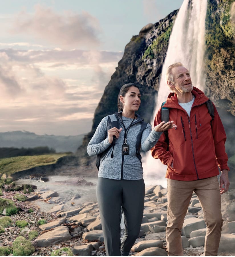 A woman and a man in hiking jackets walking on a rocky path beneath a waterfall.
