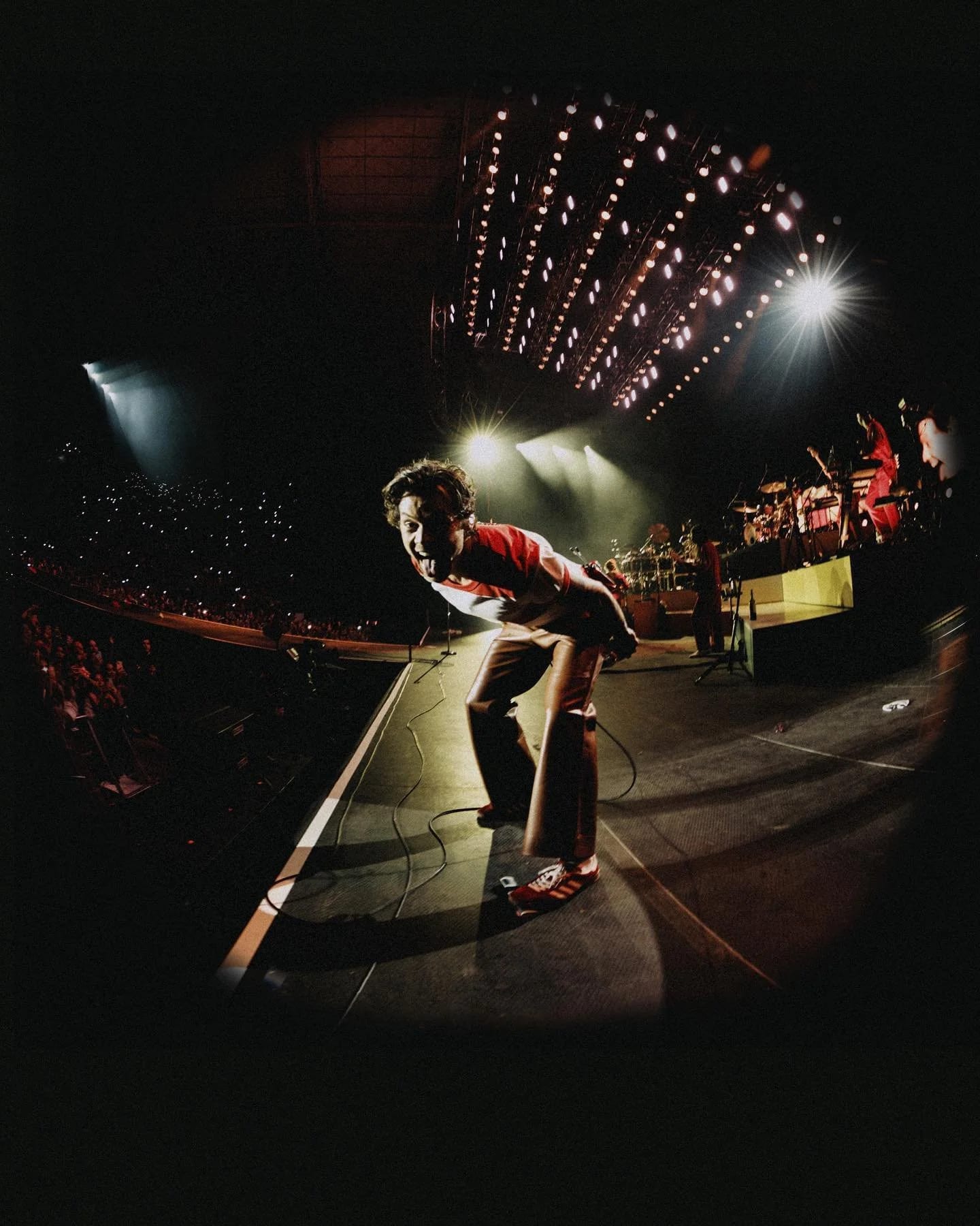 Fisheye view of Harry Styles crouching at the front of a stage before a crowd, a rig of lights overhead.