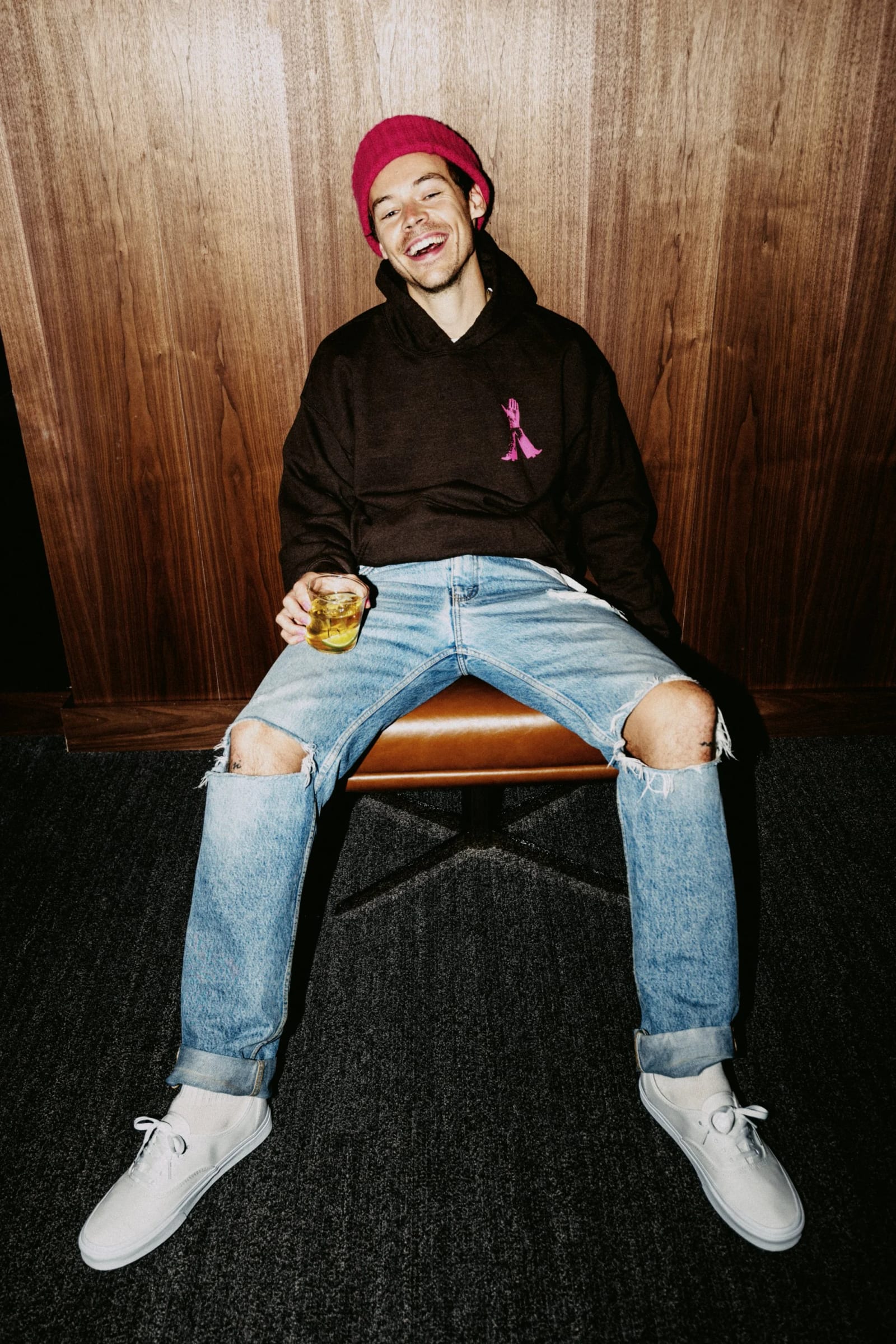 A young man in a red beanie and black hoodie sitting on a chair with his legs spread, a drink in hand, against wood panelling.