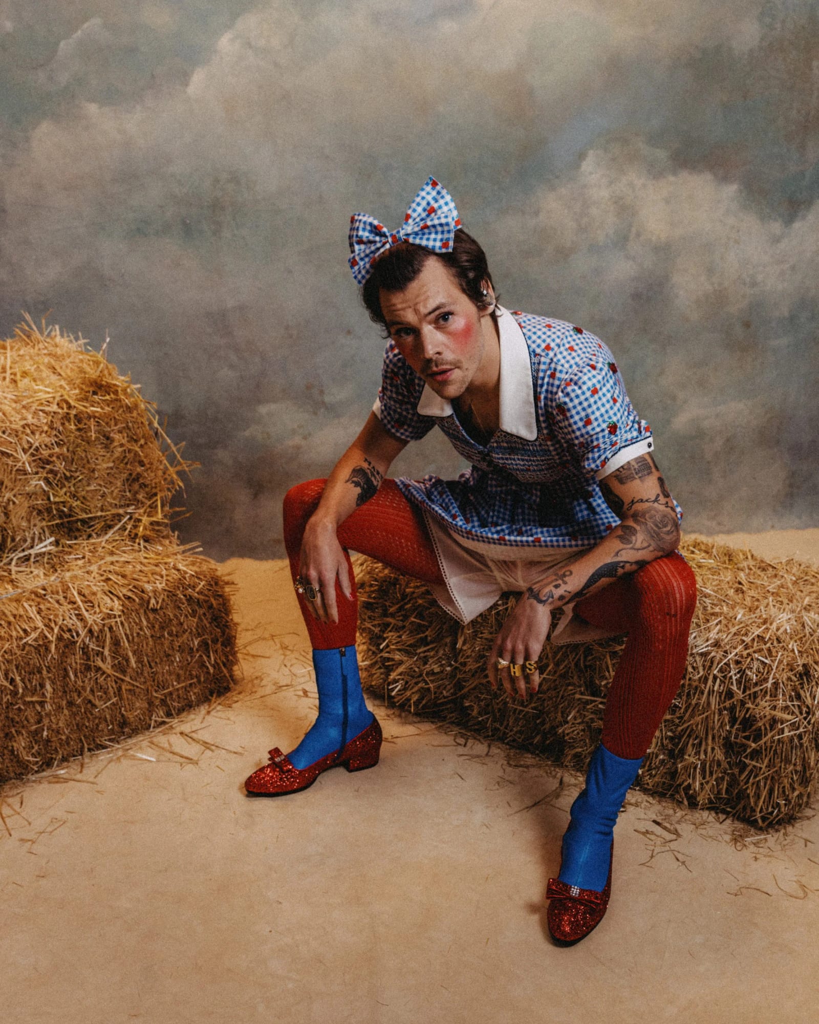 Harry Styles in a gingham dress and blue hair bow sitting on a hay bale in red tights and ruby slippers against a painted cloud backdrop.