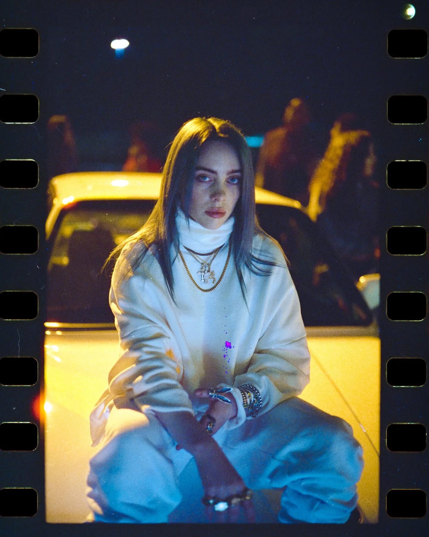 A film frame with sprocket holes at both edges: a young woman in a white turtleneck and sweatshirt crouching in front of a yellow car at night, figures standing behind her.