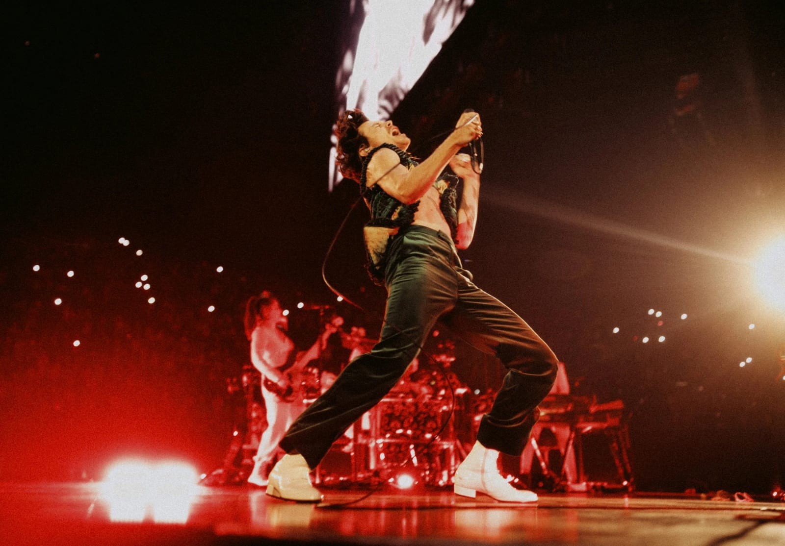 Harry Styles bent over a microphone stand on a red-lit stage, his band behind him.