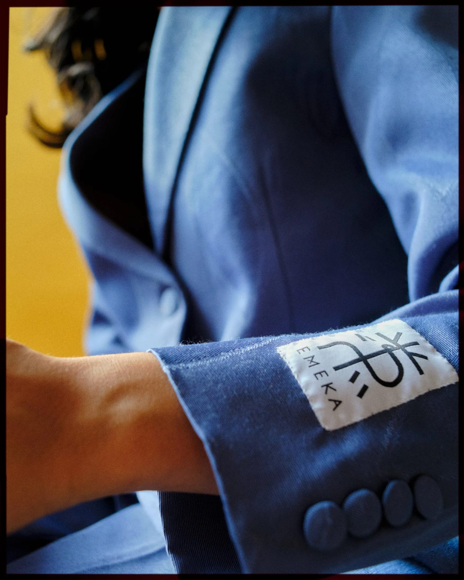 Close-up of a blue jacket sleeve with a white EMEKA label on the cuff, against a yellow wall.